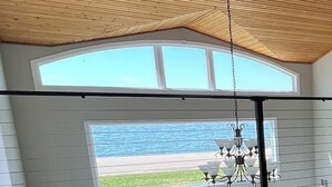A room with a large window overlooking a beach, a white table, a chair, and a hanging light fixture.