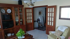 Ein Wohnzimmer mit einem Holzkasten, einem Fernseher, einer Zimmerpflanze, einem gemusterten Teppich und einem cremefarbenen Sessel.
