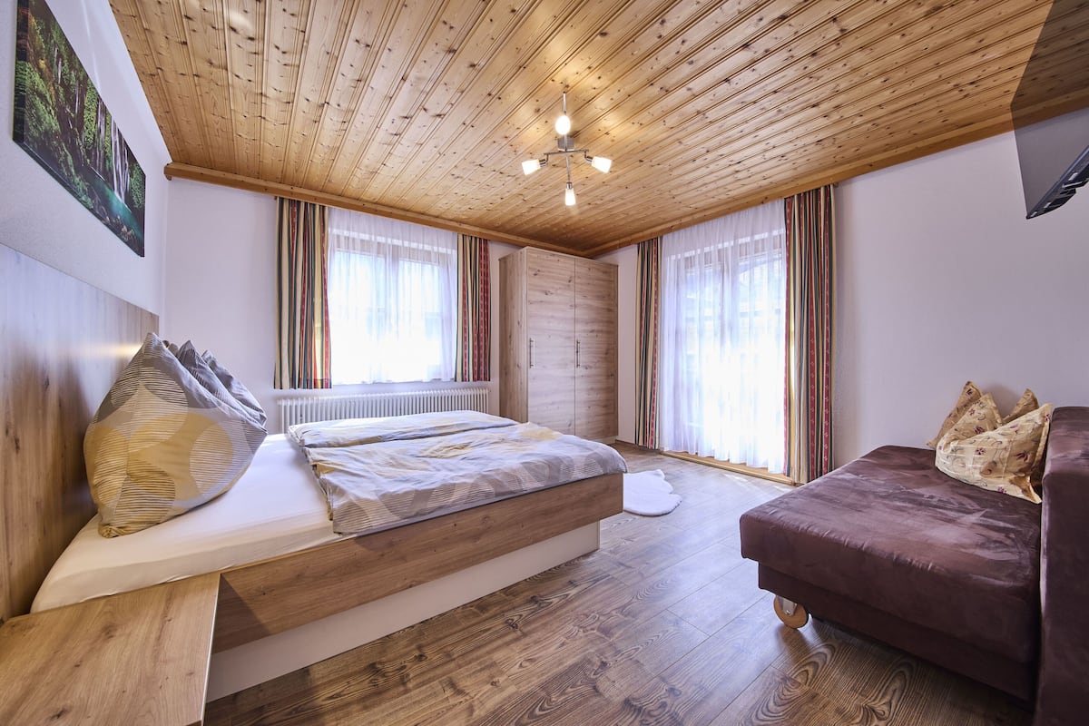 A bedroom with a wooden ceiling, a bed with a patterned bedspread, a brown sofa, and a radiator.