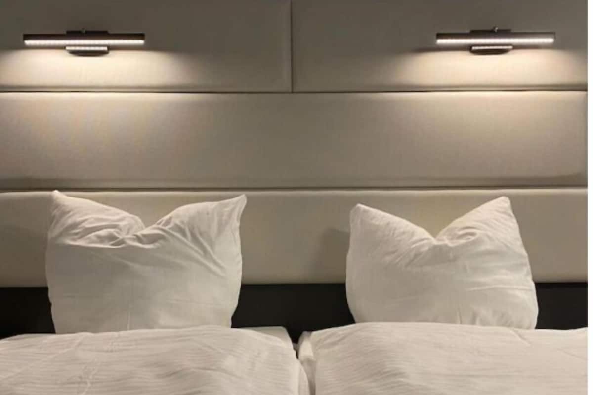 Un lit avec du linge de lit blanc et des oreillers, une tête de lit dotée d’un éclairage intégré et un mur aux couleurs neutres.