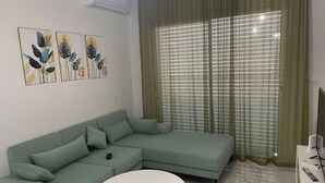 Ein modernes Wohnzimmer mit einem hellgrünen Ecksofa, einem runden weißen Couchtisch und einem großen Fenster mit Jalousien.