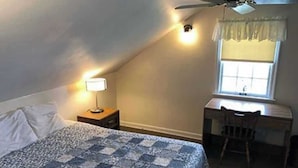 A bedroom with a bed, a desk, a chair, a window with curtains, a ceiling fan, and a nightstand with a lamp.