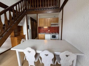 A dining area with a white table and chairs, a wooden staircase, and a kitchen with red tiles and wooden cabinets.