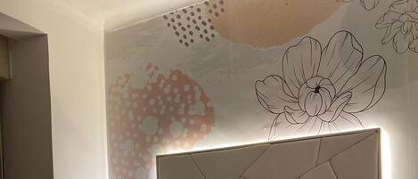 Un dormitorio con cama, mesita de noche, pared con motivos florales y geométricos, e iluminación en el techo.