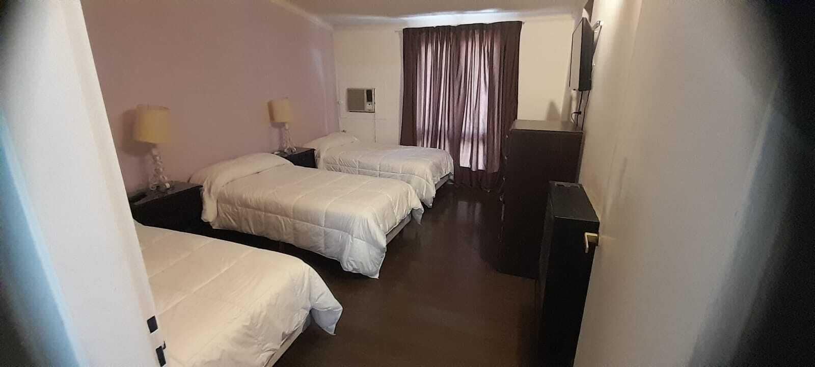 Ett hotellrum med tre sängar, en TV, ett skrivbord och ett fönster med gardiner.