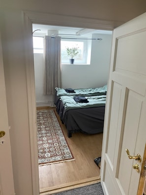 A narrow hallway leading to a bedroom with a bed, a window with a plant, and a door with a brass handle. - Herregårdshuset AS (Ullensaker)