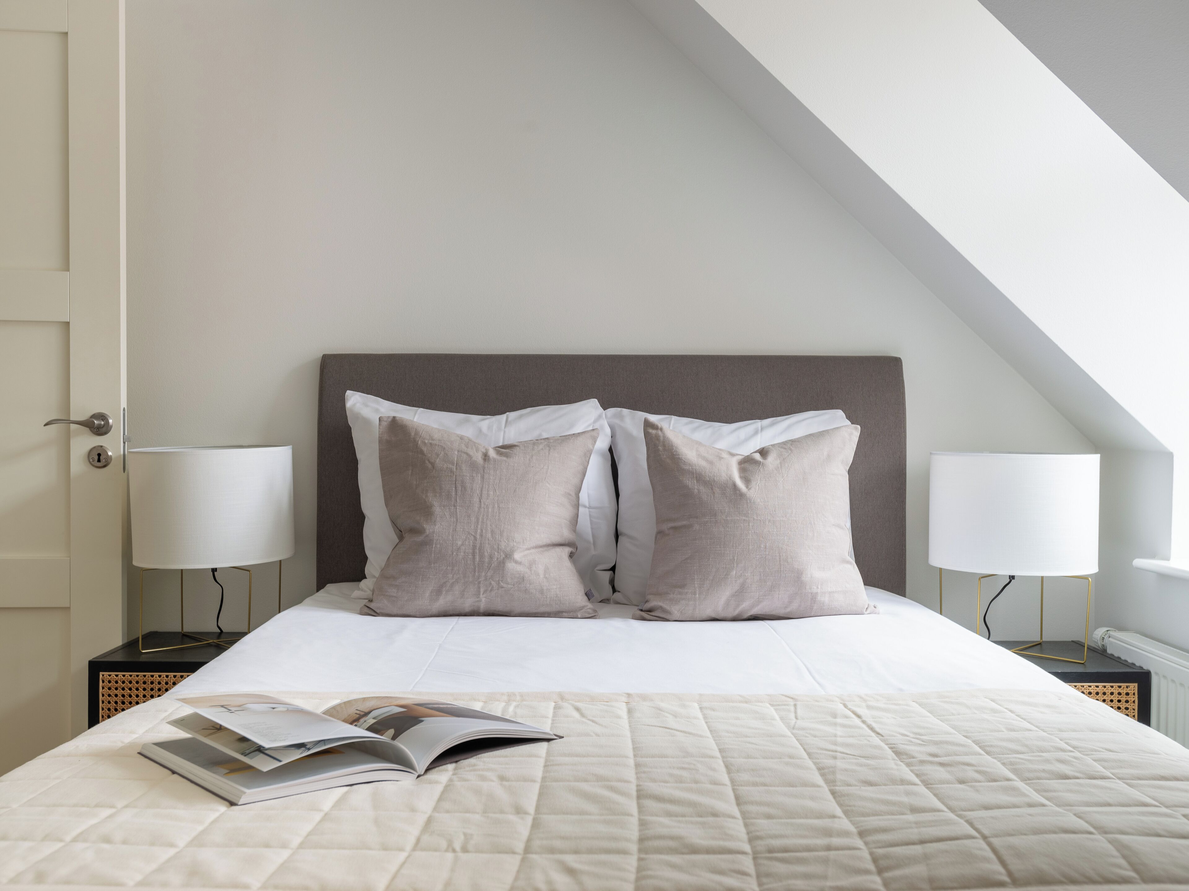Un lit bien fait, avec une couette blanche matelassée, deux oreillers et un magazine posé sur le lit. Il y a deux tables de chevet avec des lampes et une porte sur la gauche.