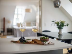 A dining table with two glasses of white wine, a wooden board with grapes, olives, and nuts, and a small bowl of green olives.
