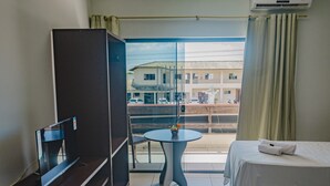 Habitación de hotel con cama, una mesita con planta en maceta, televisor sobre un mueble bajo y balcón con vista a edificios y coches estacionados.