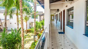 Un pasillo de hotel con suelo de baldosas, un balcón con barandilla y vistas a un patio con palmeras y plantas.