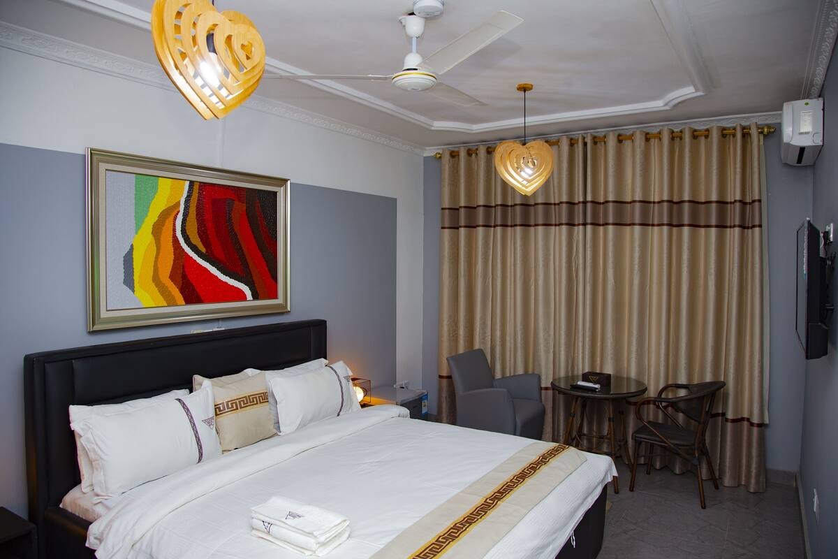 A hotel room with a bed, a desk, two chairs, a ceiling fan, and a painting on the wall.
