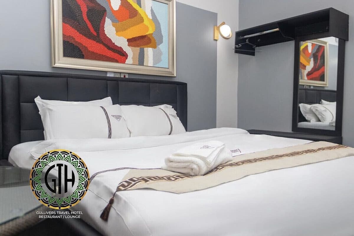 A hotel room with a bed, a headboard, a wall-mounted light, a painting, and a logo.