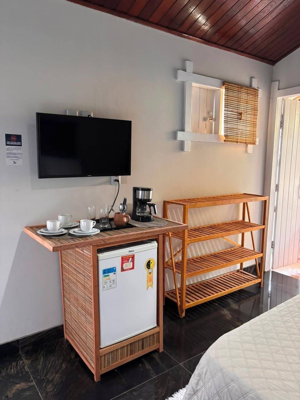 A hotel room with a flat-screen TV mounted on the wall, a small kitchenette with a refrigerator, a coffee maker, and a wooden shelf unit.