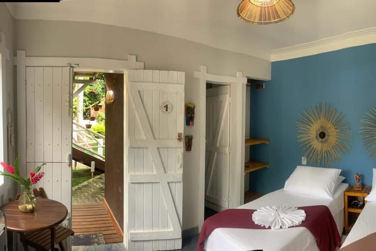 Ein Schlafzimmer mit zwei Betten, einem Holztisch mit einer Vase und Blick auf einen Garten durch offene Türen.