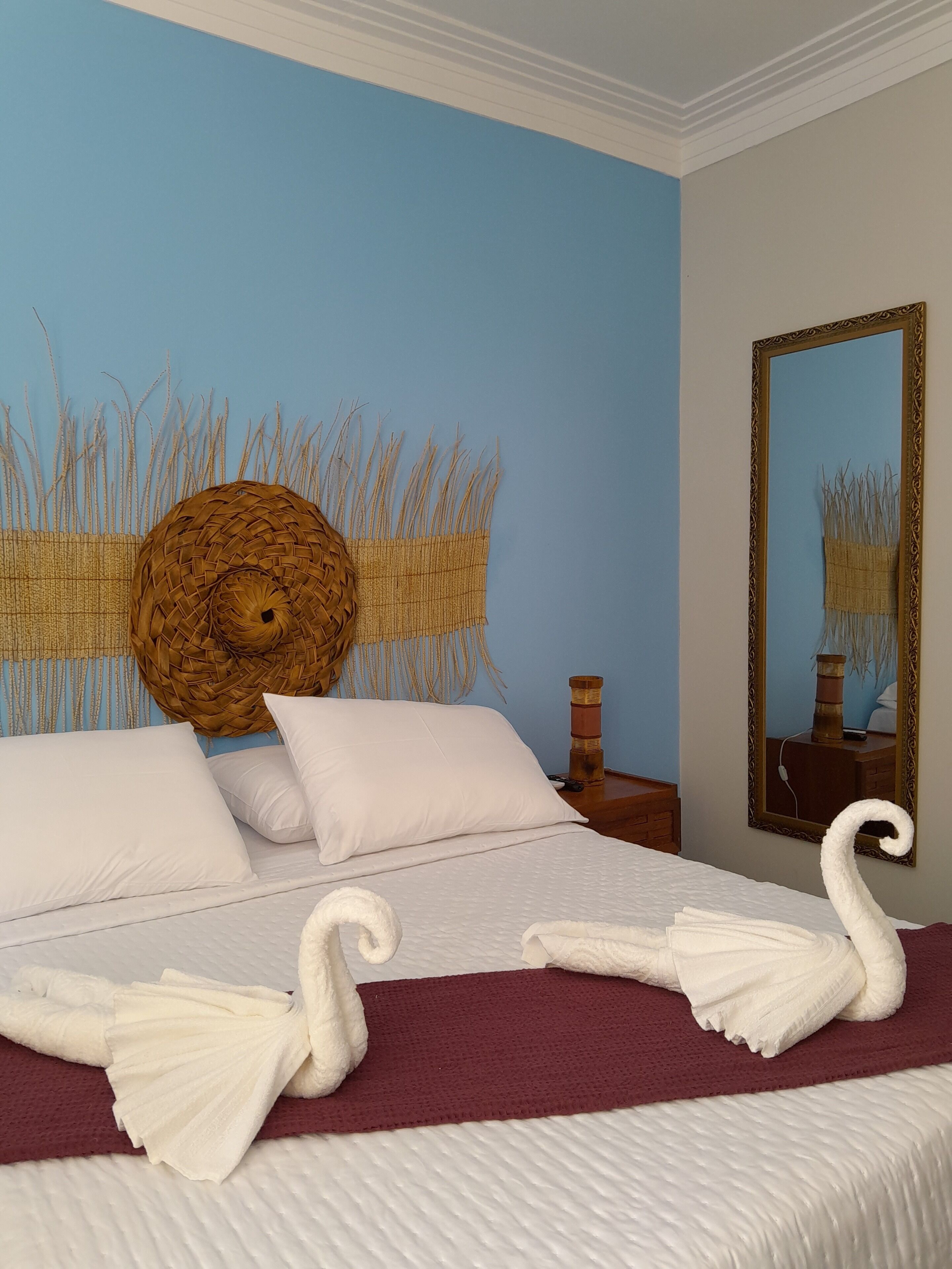 A neatly made bed with white linens and decorative swan pillows, a woven wall hanging, and a mirror with a decorative frame.