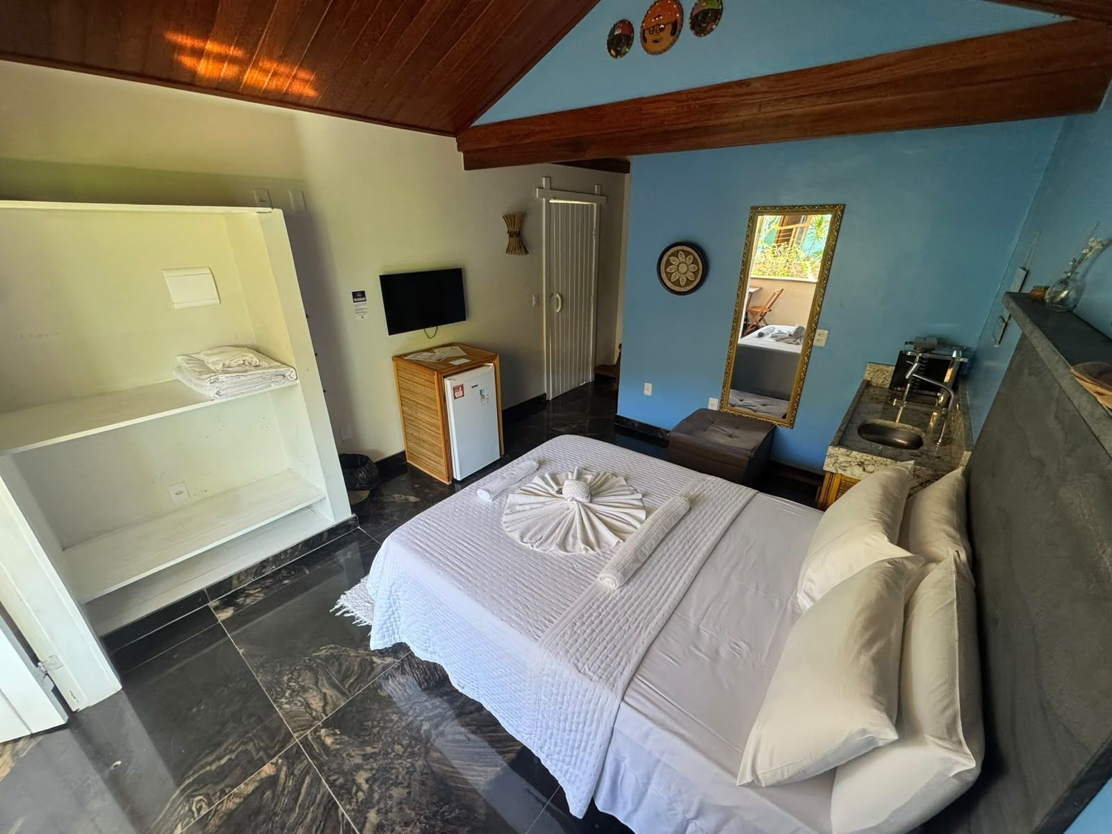A hotel room with a bed, a TV, a small refrigerator, a wooden cabinet, a mirror, and a bathroom with a sink and toilet.