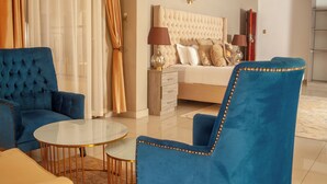 Ein modernes Hotelzimmer mit einem gesteppten Bettvorleger, einem blauen Sessel, einem runden Glastisch und einem beigefarbenen Sofa.