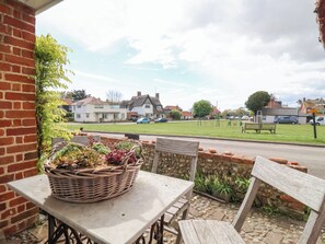 A patio with a table and chairs, a wicker basket with plants, and a view of houses and a road. - Beta Cottage (Reydon)