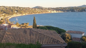 Vista costera con tejados de teja terracota, una playa y un paisaje montañoso al fondo.
