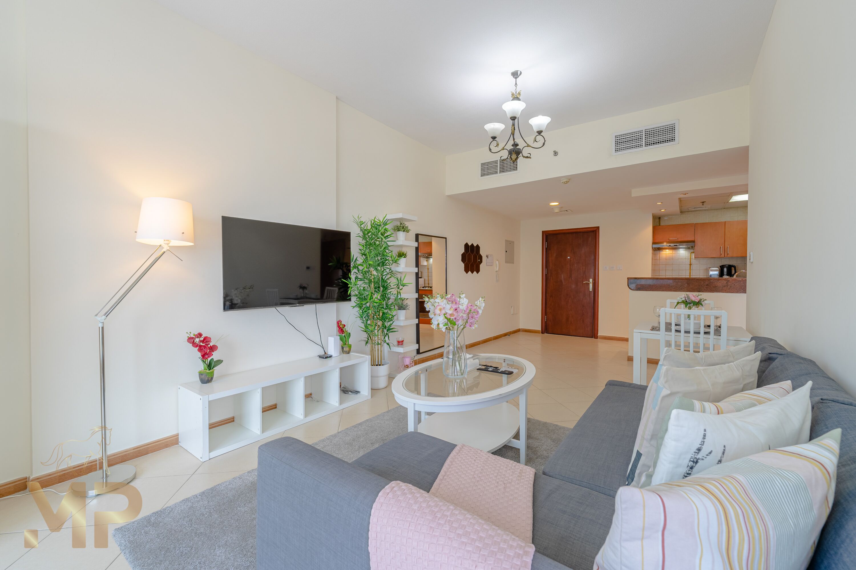 Una sala moderna con un sofá gris, una mesa de centro blanca, un televisor de pantalla plana, una lámpara de pie y una planta en maceta.
