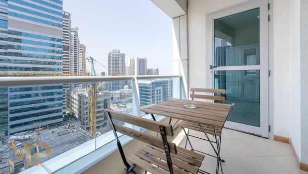A balcony with a wooden table and chairs, overlooking a cityscape with modern buildings and construction cranes.