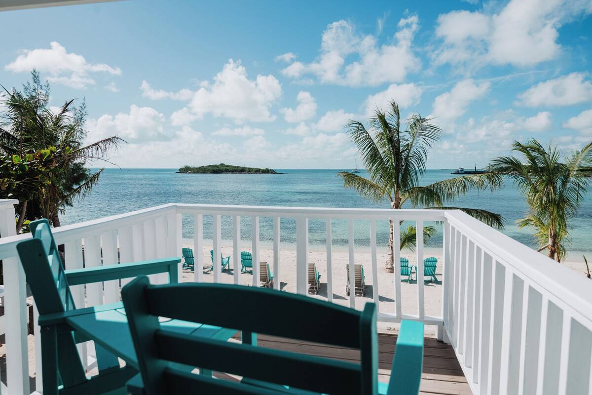 A wooden deck with green chairs overlooking a beach with clear blue water and a small island in the distance.