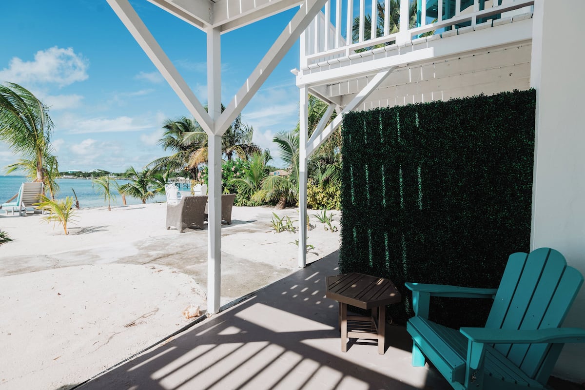 A beachside patio with a white pergola, a wooden table, and two turquoise Adirondack chairs.