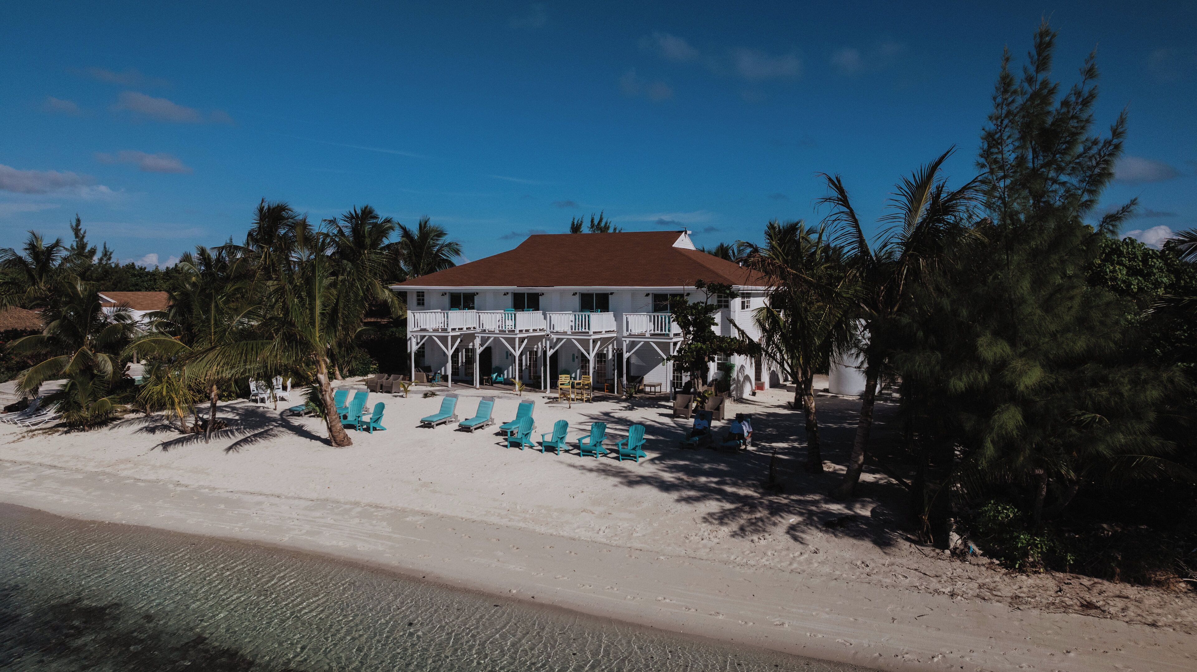 A beachfront property with a white building featuring a balcony, surrounded by palm trees and beach chairs.