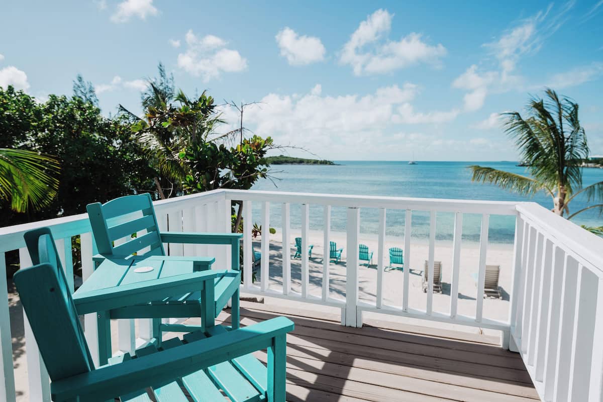 A wooden deck with teal chairs and a table, overlooking a beach with lounge chairs and palm trees.