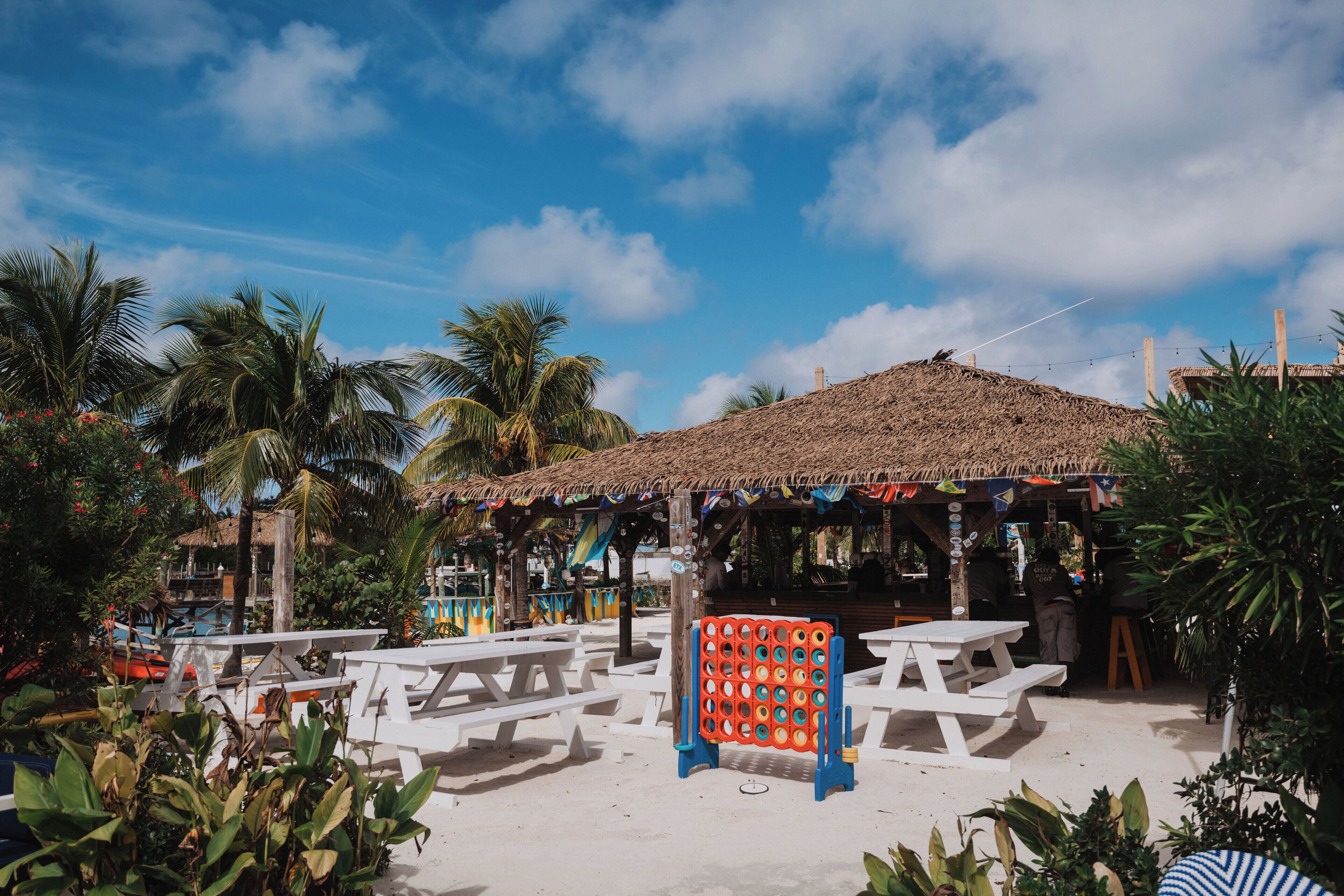A beachside area with white picnic tables, a thatched-roof structure, and palm trees.
