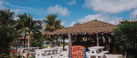 A beachside area with white picnic tables, a thatched-roof structure, and palm trees.