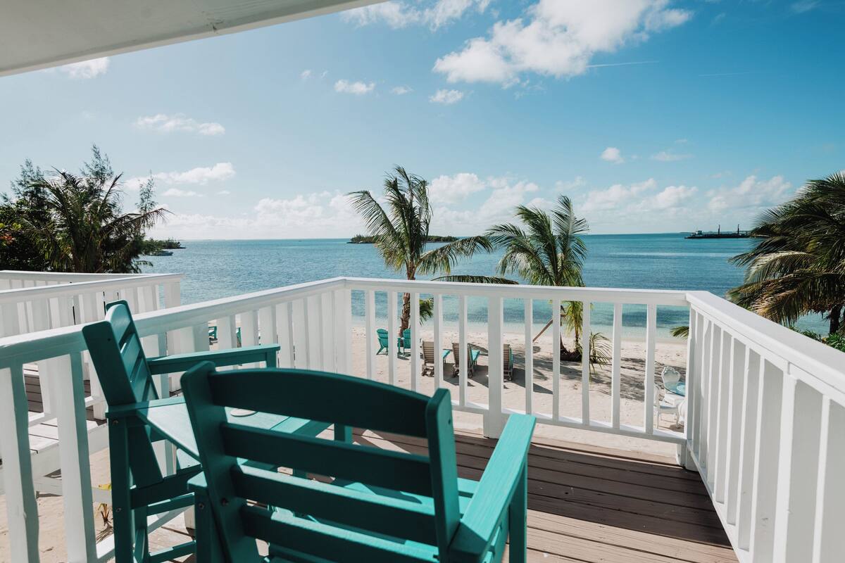 A wooden deck with white railings overlooking a beach with palm trees and a clear blue sky.