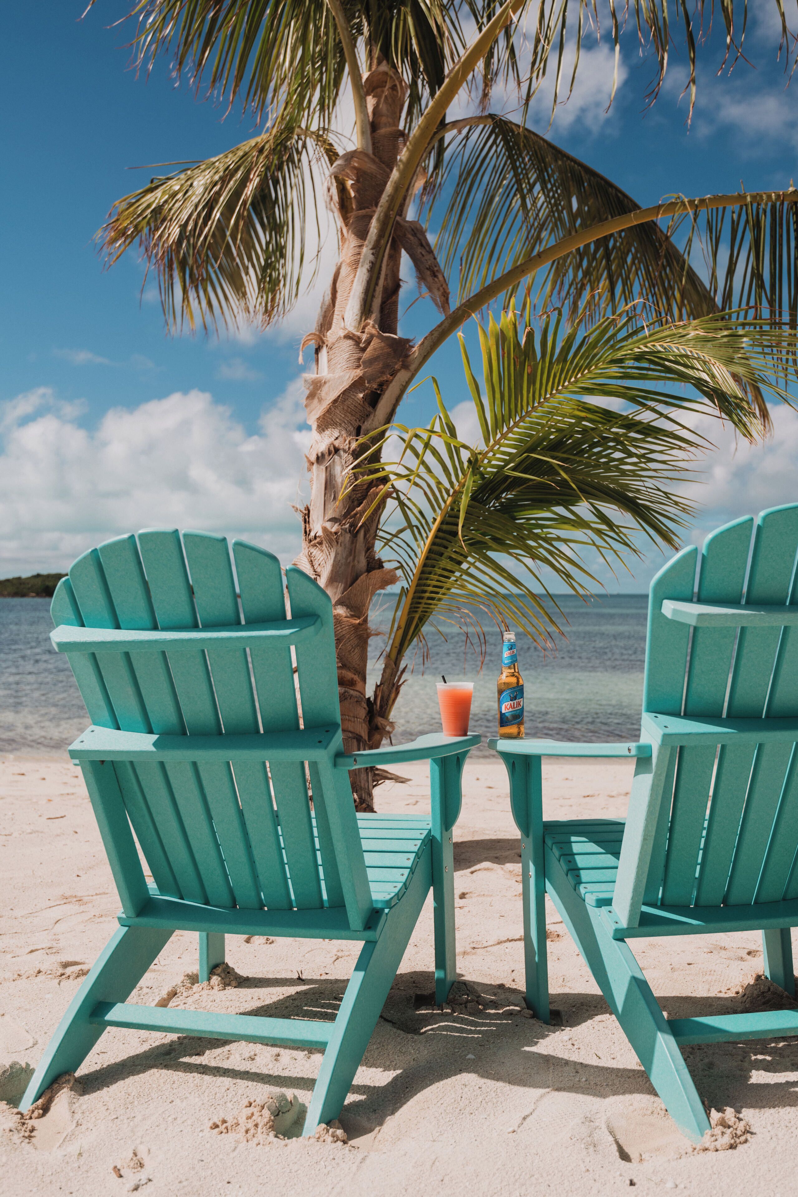 Zwei türkisblaue Adirondack-Stühle an einem Sandstrand, mit einer Palme und einer Bierflasche auf einem kleinen Tisch.