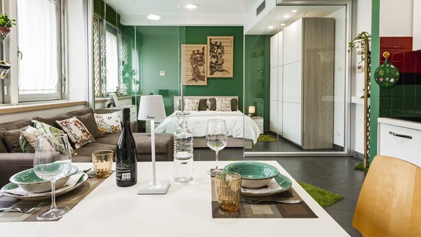 Une chambre d’hôtel moderne dotée d’un coin repas, d’un lit avec un mur vert et d’une cuisine visible à travers une porte ouverte.