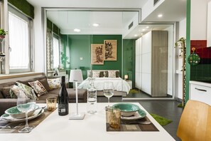 Ein modernes Hotelzimmer mit Essbereich, einem Bett an einer grünen Wand und einer durchgehenden Tür erkennbaren Küche.