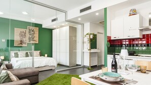Une chambre d’hôtel moderne comprenant un lit, une table à manger avec des chaises, un coin cuisine avec des placards et un évier, ainsi qu’un mur végétal agrémenté d’œuvres d’art.