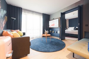 Ein modernes Hotelzimmer mit Sofa, einem runden Teppich, einem kleinen Tisch mit Pflanze, einem Bett und einem großen Fenster mit Vorhängen.