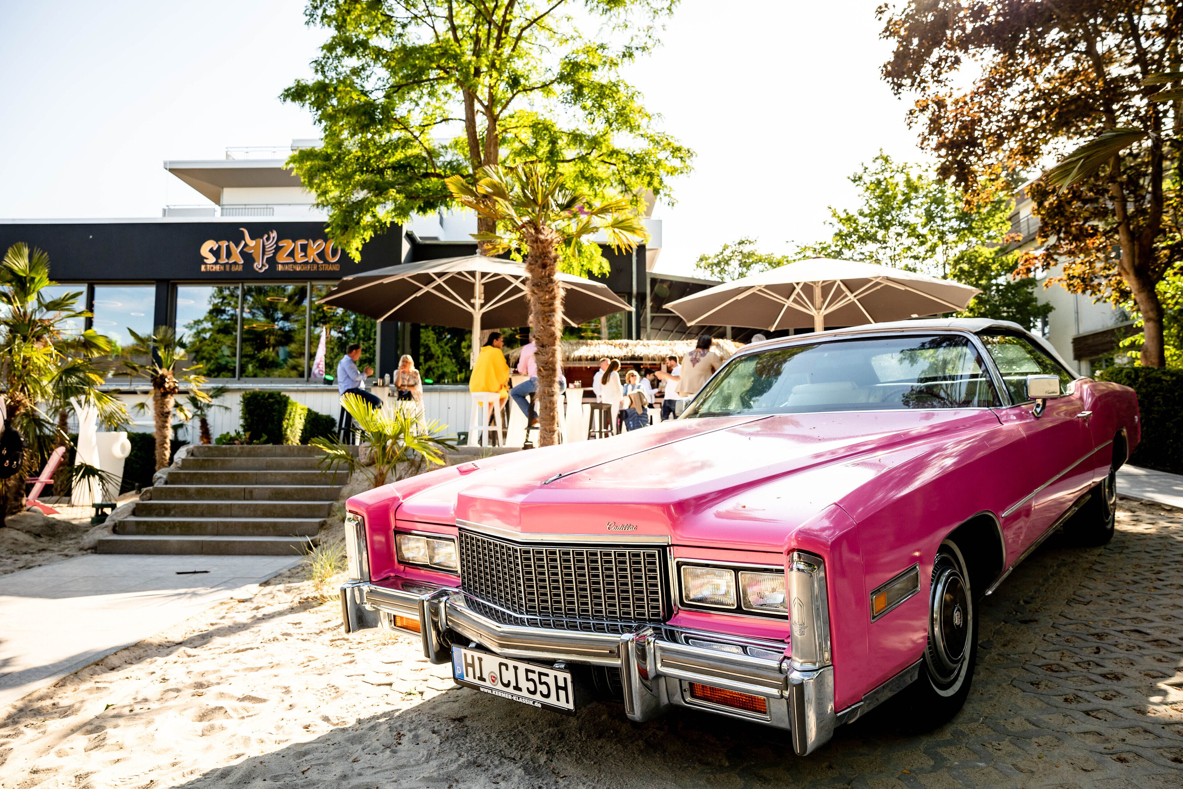Un Cadillac rosa antiguo estacionado frente a un edificio moderno con mesas al aire libre.