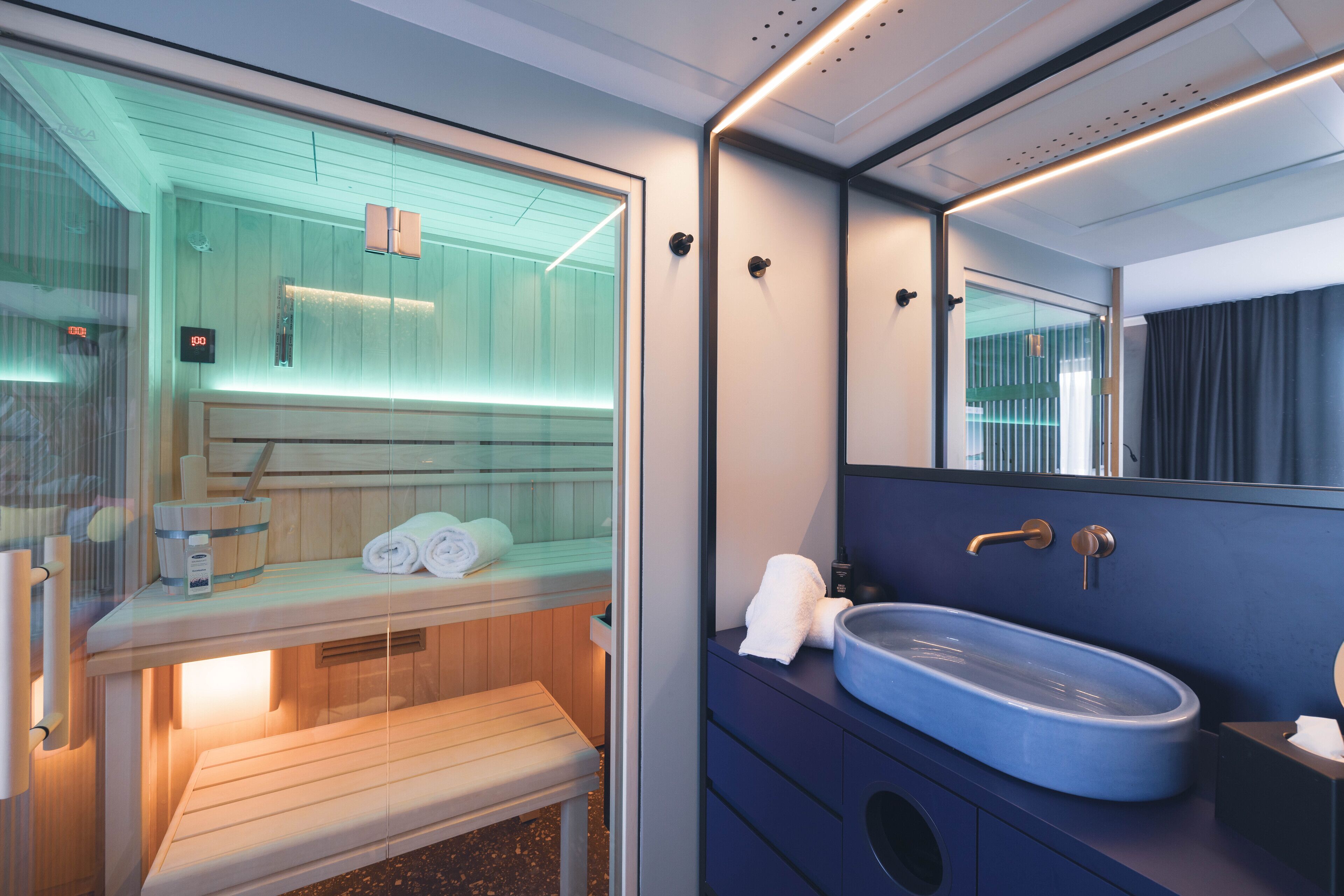 Une salle de bain moderne avec un sauna en verre, un banc en bois, un grand lavabo bleu et un miroir.