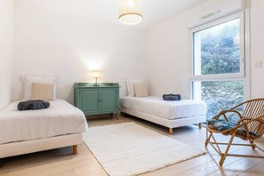 A bedroom with two beds, a green cabinet, a wicker chair, and a large window.