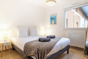 A bedroom with a bed, a bedside table with a lamp, a framed picture on the wall, and a window with a view of a building.