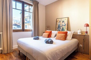 Hotellihuone, jossa on suuri sänky, ikkuna, jossa on verhot, seinällä taulu ja yöpöytä, jossa on lamppu.