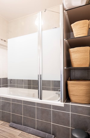A bathroom with a glass-enclosed bathtub, dark grey tiles, and wicker baskets on shelves.