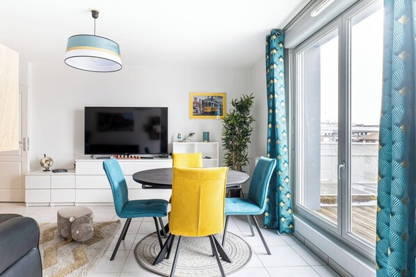 A modern living room with a TV, dining table, chairs, and a large window with curtains.