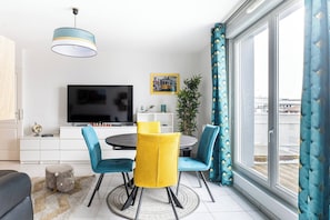A modern living room with a TV, dining table, chairs, and a large window with curtains.