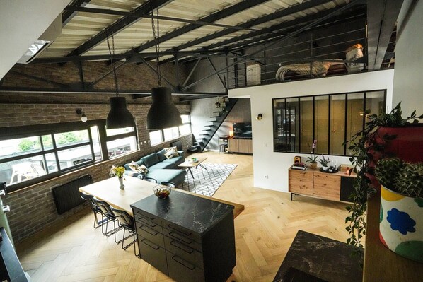 A spacious loft with a high ceiling, exposed brick walls, a dining area with a large island, and a staircase leading to an upper level.