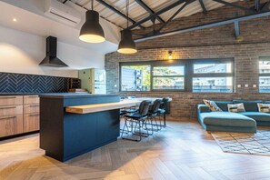 A modern kitchen with a central island, bar stools, and a sofa in a living area with large windows.