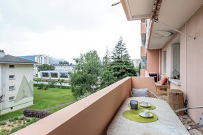 En balkon med et bord til to, en fletkurv og udsigt over en grøn græsplæne og bygninger.