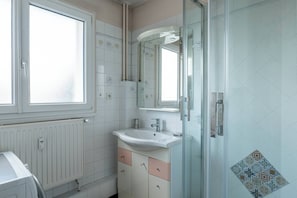 A bathroom with a window, a sink, a mirror, a shower, and a radiator.