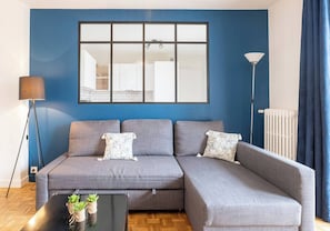 A modern living room with a grey sectional sofa, a black coffee table with plants, a floor lamp, and a large window with a view of the kitchen.